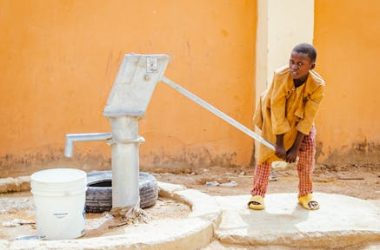 pexels-photo-32154739-32154739 Young child using a manual water pump in an African village to draw water into a bucket.
